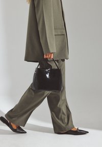 Persona caminando con un traje verde oliva, llevando un bolso negro y calzando zapatos negros de punta con tira trasera, sobre un fondo liso.