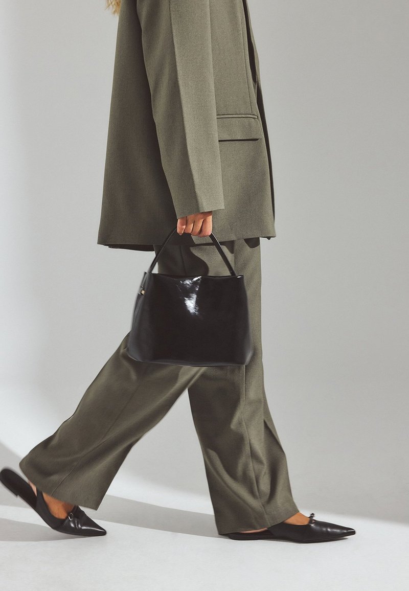 Persona caminando con un traje verde oliva, llevando un bolso negro y calzando zapatos negros de punta con tira trasera, sobre un fondo liso.