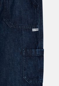 Tmavě modré denimové cargo kalhoty s náprsní kapsou a štítkem s nápisem "HILFIGER." Obsahují zesílené švy a texturovaný vzhled látky.
