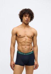 Mišićavi muškarac s kovrčavom kosom nosi tamnoplave Tommy Hilfiger bokserice s zelenim pojasom, stoji nasuprot jednostavne bijele pozadine.