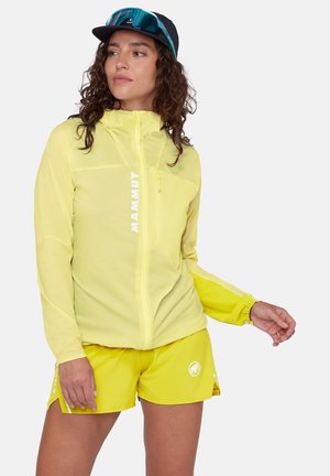 Frau trägt gelbe Mammut-Jacke und Shorts mit Cap und Sonnenbrille, blickt seitlich vor einfarbigem Hintergrund.