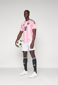 Lyserød fodboldtrøje med sorte accenter og logo, parret med hvide shorts og sorte sokker med striber, som holder en fodbold.