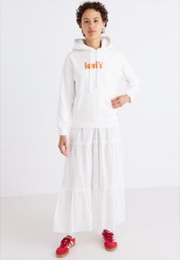 Vit hoodtröja med orange "Levi's"-logotyp, ihopparad med en vit lager-på-lager maxikjol. Modellen bär röda sneakers. Bakgrunden är enfärgad.