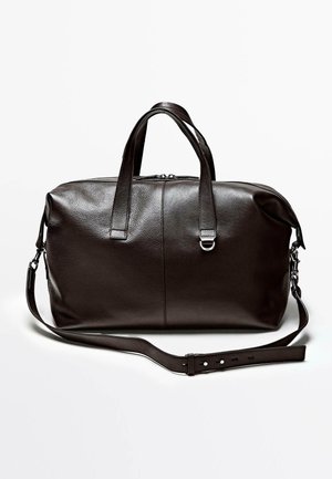 Ciemnobrązowa skórzana torba podróżna z podwójnymi uchwytami i regulowanym paskiem na ramię na białym tle.