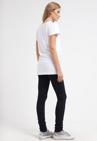 T-shirt branco de manga curta combinado com jeans skinny escuros e sapatilhas cinzentas, visto de lado. Tecido suave, design simples e corte clássico.