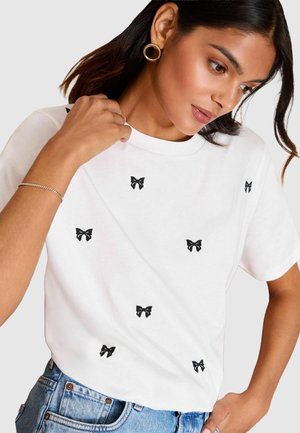 T-shirt en coton blanc avec des broderies de nœuds noirs éparpillées, un col rond et des manches courtes, associé à un short en denim bleu.