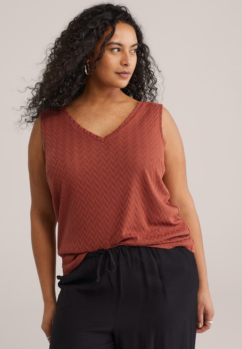 Femme aux cheveux bouclés portant un haut sans manches couleur rouille avec motif chevron et un pantalon noir à cordon, regardant sur le côté.