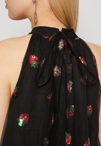 Svart genomskinlig topp med röd och grön paljettbroderi av jordgubbar, med hög hals och knytning i ryggen. Texturerat tyg med lätt glans.