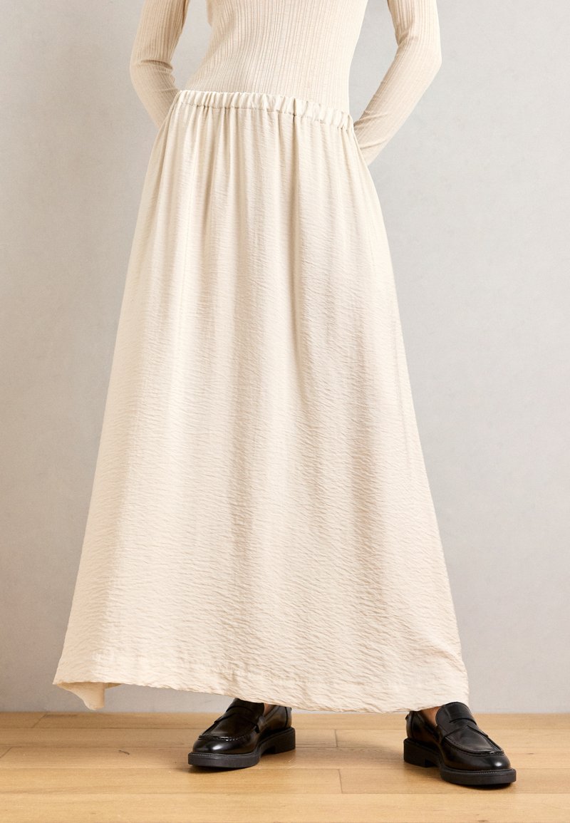 Cream-colored maxi skirt with a textured finish and elastic waistband; paired with black loafers, set against a neutral background.