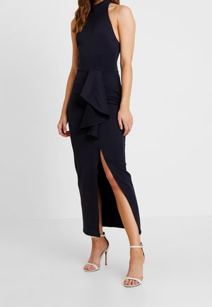 Vrouw draagt een mouwloze zwarte jurk met een hoge hals, een volantdetail en een hoge split aan de dij, gecombineerd met witte sandalen met hoge hakken.