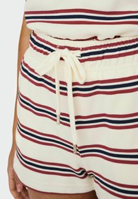 Pantaloni corti a righe con vita elastica e chiusura con coulisse. I colori includono crema, rosso e blu navy. Tessuto a trama con righe orizzontali.