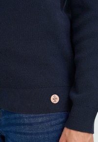 Pull en tricot bleu marine avec des bords côtelés aux poignets et à l'ourlet. Présente une petite étiquette logo beige sur le côté inférieur. Texture douce, design décontracté.