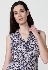 Marineblauwe mouwloze blouse met een knoopsluiting aan de voorkant, voorzien van roze en crème bloemenpatronen en een kraagnek. Gladde stoffen textuur.