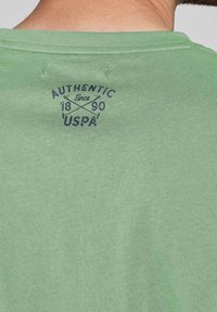 Zelené bavlněné tričko s kulatým výstřihem, krátkými rukávy a námořnicky modrým grafickým potiskem "AUTHENTIC since 1890 USPA" na zádech.