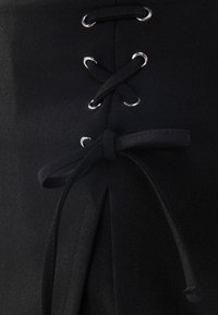 Tejido negro con detalle de encaje de ojal y un lazo. Textura suave, con acentos de hardware plateado alrededor del encaje para añadir interés visual.
