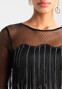 Haut en maille noire à texture pointillée, avec des détails de franges le long de l'ourlet et des bandes argentées, fini avec un col rond.