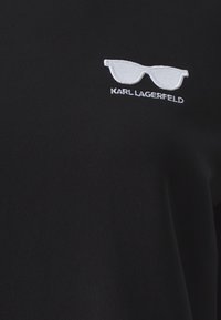 Svart hettegenser med hvit broderte solbriller og teksten "KARL LAGERFELD." Glatt stoff med en ensfarget tekstur og en avslappet design.