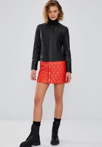 Zwarte leren jas met gequiltte schouders, ritssluiting en twee zijzakken; gecombineerd met een rode gequiltte rok met drukknoopsluiting.
