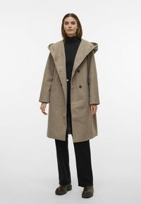 Cappotto in misto lana marrone con un ampio cappuccio, doppio petto anteriore e tasche laterali, abbinato a pantaloni neri e scarpe marroni chunky.