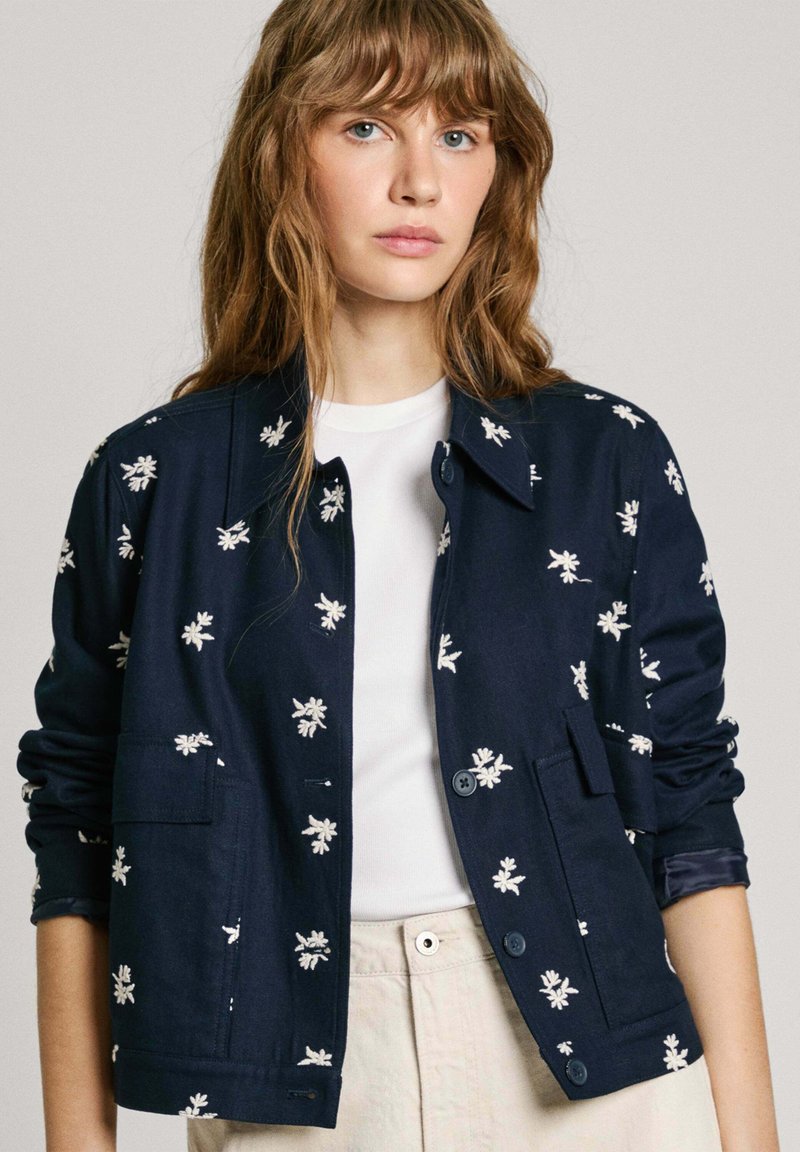 Veste bleu marine en tissu avec broderies florales blanches, dotée d'un col, d'une fermeture à boutons et de deux poches avant.
