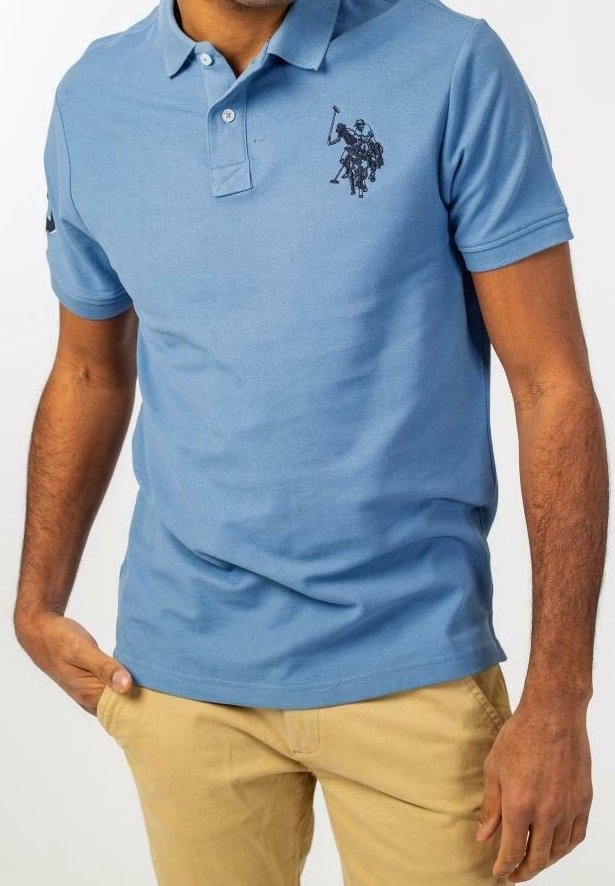 Homme portant un polo bleu clair avec un logo de joueur de polo noir et un pantalon beige, debout avec une main dans la poche.