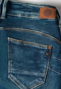 Un jean en denim bleu foncé présente une finition texturée, des coutures contrastées, une étiquette en cuir à la taille et une poche arrière avec une étiquette de marque.