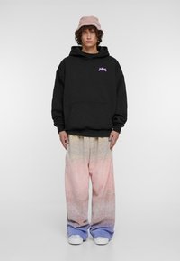 Juodas džemperis su violetiniu grafiku, didelė priekinė kišenė, derinamas su per didelėmis sportinėmis kelnėmis, kurios turi rožinį ir mėlyną gradientą, ir baltomis sportbačiais.