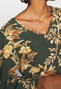 Blusa floreale con scollatura a V profonda, realizzata in tessuto verde decorato con fiori gialli e crema, texture liscia e maniche lunghe.