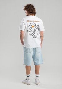 Camiseta blanca de algodón con un diseño en negro y naranja de Keith Haring que presenta una flor y una figura en la parte posterior. Corte casual con mangas cortas.