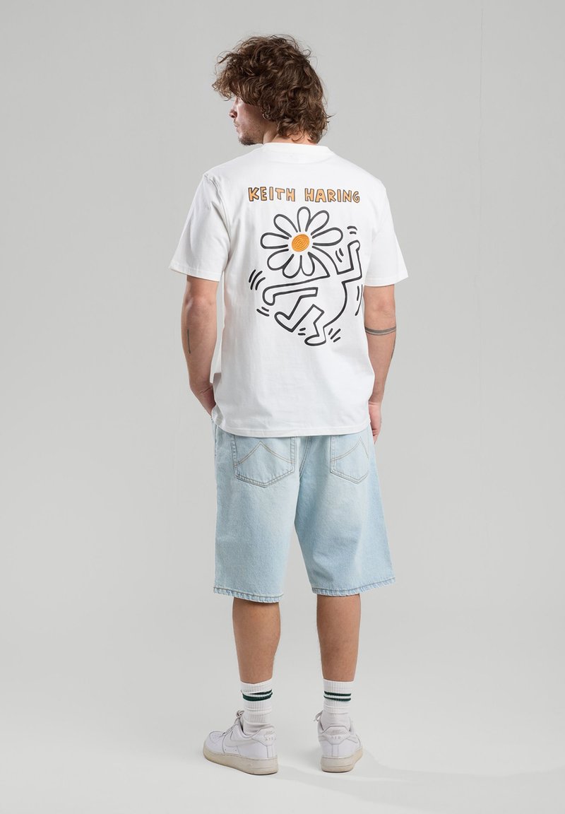 Camiseta blanca de algodón con un diseño en negro y naranja de Keith Haring que presenta una flor y una figura en la parte posterior. Corte casual con mangas cortas.