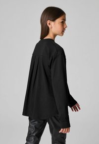 Musthave musta pikkade varrukatega shirt, millel on lõdva lõikega seljaosa ja dekoratiivsed aktsendid varrukaotstel. Koos läikivate mustade pükstega.
