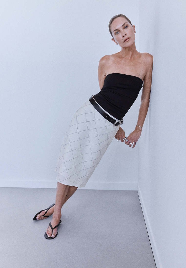 Femme en haut noir sans bretelles, jupe blanche à carreaux et sandales noires, appuyée contre un mur blanc dans une pièce minimaliste.