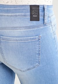 Světle modré džíny z denimu s detailním pohledem na pás, obsahující černou nálepku s nápisem "ULTRA MOVE" a viditelnými detaily šití.