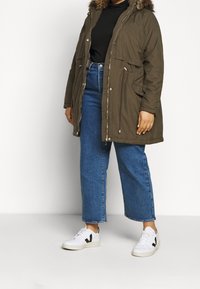 Parka verde oliva con capucha de pelo sintético, cierre de cremallera y botones a presión, y bolsillos frontales. Combinado con jeans de pierna ancha azules y zapatillas blancas.