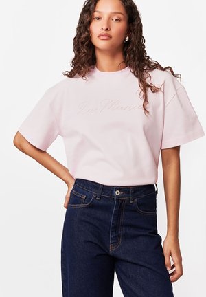 Młoda kobieta z kręconymi włosami, ubrana w jasnoróżową koszulkę z krótkim rękawem i ciemnoniebieskie, wysokie spodnie dżinsowe, pozująca z jedną ręką na biodrze.