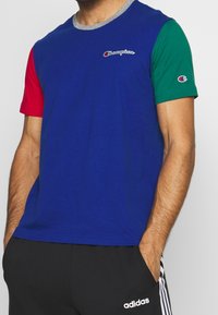 Muž v modrém tričku Champion s červenými a zelenými rukávy a černých kalhotách Adidas s bílými pruhy a logem, s rukama v kapsách.