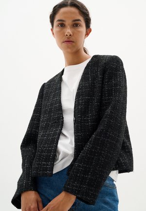 Black textured jacket with open front, long sleeves, and a loose fit. Worn over a white shirt and paired with blue jeans.