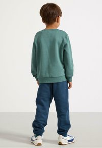 Felpa verde con polsini e orlo a costine, abbinata a pantaloni della tuta blu scuro. Le calzature presentano una base bianca con accenti blu e gialli.