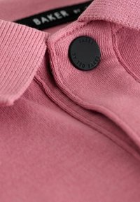 Nahaufnahme eines pinken Strickstoffs mit einem schwarzen Knopf und einem mit "Baker by Ted Baker" geprägten Etikett.