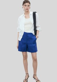 Modré ľanové šortky so štruktúrovaným strihom, v kombinácii s bielym tíčkom a ľahkou, otvorenou bielou košeľou. Look dopĺňajú sandále na gumovej podrážke.