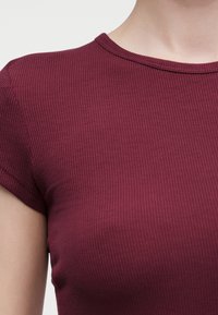 Κοντινό πλάνο ενός ατόμου που φοράει εφαρμοστό, κοντομάνικο, ριμπ πουκάμισο μπορντό με στρογγυλή λαιμόκοψη σε απλό φόντο.