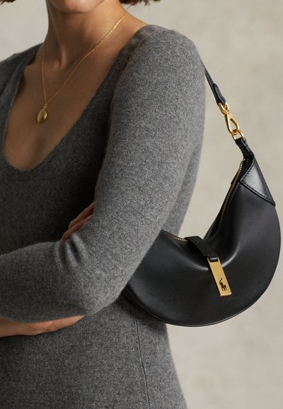 Žena v sivom tričku s dlhým rukávom, nosiaci zlatý prívesok na náhrdelníku a nesúci čiernu koženú kabelku v tvare polmesiaca so zlatými kovovými prvkami.