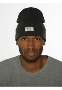 Bonnet en tricot gris avec un revers replié et une patch rectangulaire étiqueté "NXG PROTEST". Porté avec un t-shirt à col rond gris.