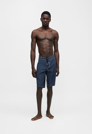 Vékony, sötét bőrű férfi áll mezítláb, tengerészkék középhosszú fürdőnadrágot visel, amelynek derékán húzózsinór van, egyszínű világos háttér előtt.