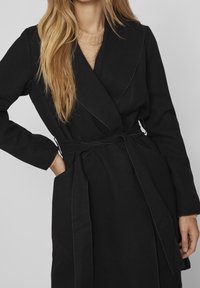 Musta käärylle takki pitkät hihat, jossa on vyötärö vedetty yhteen ja sivutaskut. Valmistettu pehmeästä, tekstuurisesta kankaasta, jossa on kontrastiväriset ommeltyöt.