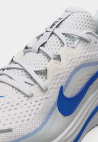 Bela tekaška copat Nike z modrim logotipom, zgornji del iz zračne mrežice in jezičkom z napisom "PHANUFLD 12".