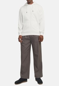 Haut à zip blanc avec capuche à cordon, associé à un pantalon gris ample. Le modèle porte des chaussures noires avec des semelles beige.