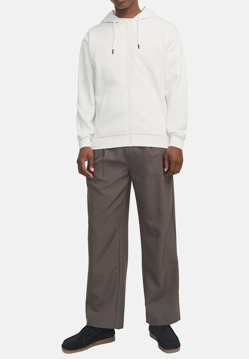 Haut à zip blanc avec capuche à cordon, associé à un pantalon gris ample. Le modèle porte des chaussures noires avec des semelles beige.