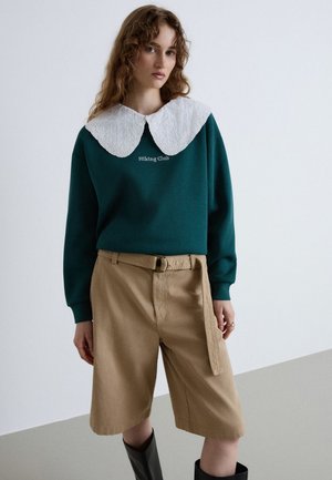 Junge Frau mit lockigem Haar, die einen grünen Pullover mit weißem Spitzenkragen, beige taillierte Shorts und schwarze knielange Stiefel trägt.