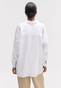 Camicia bianca di lino con colletto, maniche lunghe e spacchi laterali, caratterizzata da una vestibilità ampia e un tessuto texturizzato. Mostrata da dietro.